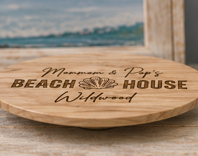 A round wooden serving board engraved with 'Mom & Pop's BEACH HOUSE Wildwood' and a seashell motif, shown on a rustic wooden surface with a blurred beach-themed background.