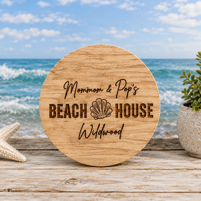 A round wooden plaque engraved with 'Mom & Pop's BEACH HOUSE Wildwood' and a seashell motif, displayed on a wooden surface with a blurred beach and ocean background.