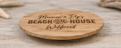 A round natural wood coaster engraved with 'Mom & Pop's BEACH HOUSE Wildwood' and a seashell motif, placed on a light wood tabletop with blurred seashells in the background.
