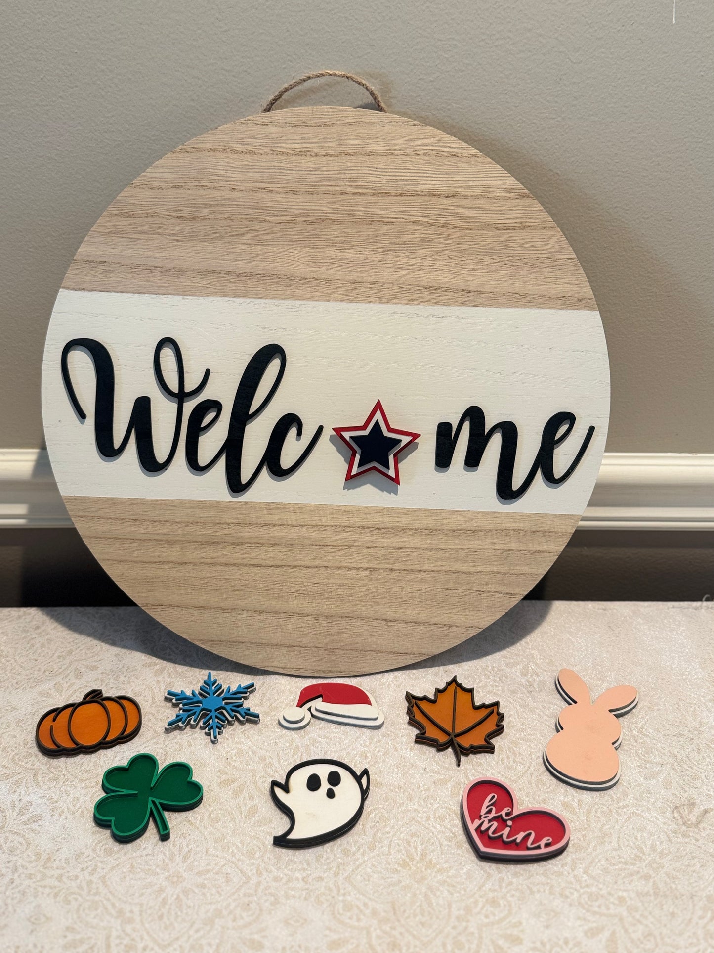 Round wooden sign with 'Welc*me' text and decorative pins on a neutral background