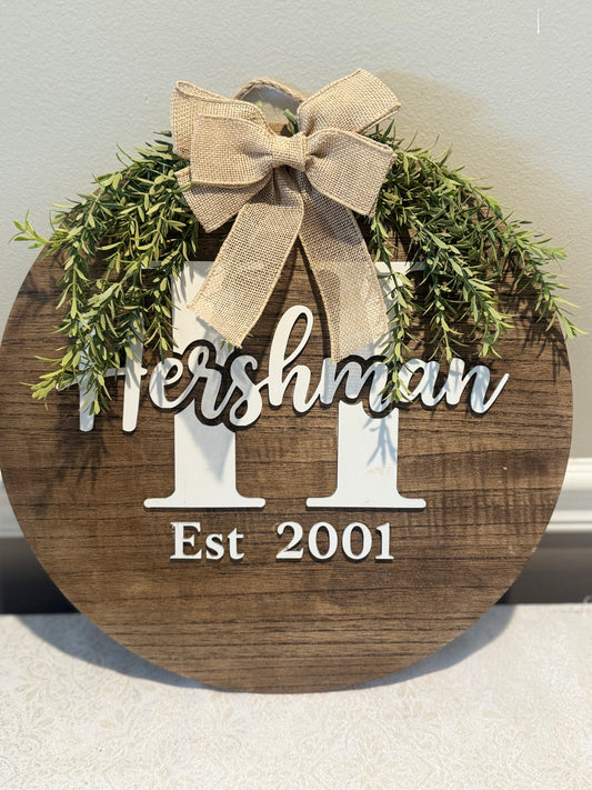 Wooden plaque with 'Hershman' and 'Est 2001' text, decorated with greenery and a bow.