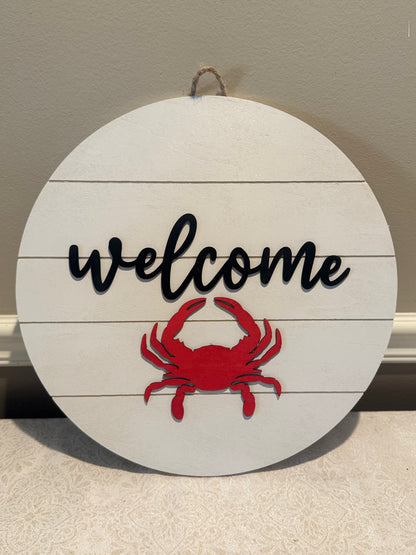 Round wooden sign with 'welcome' text and red crab design on a neutral background